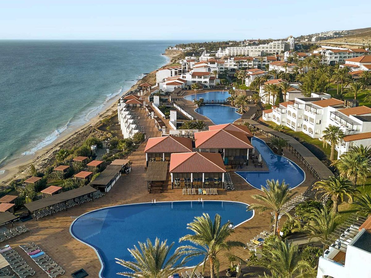 Aerial view of the resort's white low-rise blocks with terracotta roofs stepping along a clifftop above a long sandy beach, with two large freeform pools and palms in the foreground.