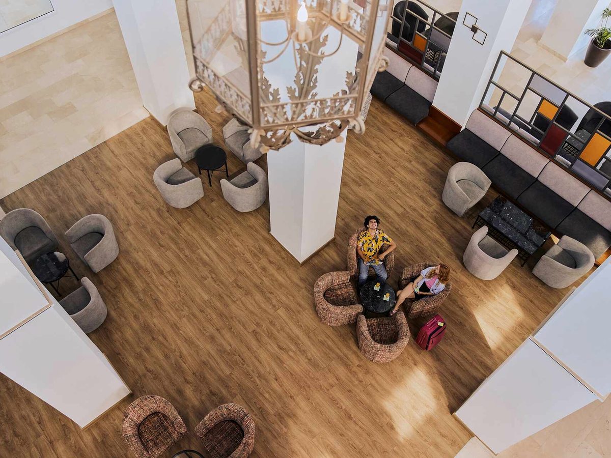 Hotel lobby seen from a balcony above, with grey armchairs and side tables on a wood-effect floor, a large pendant chandelier and guests' suitcases by the seating.