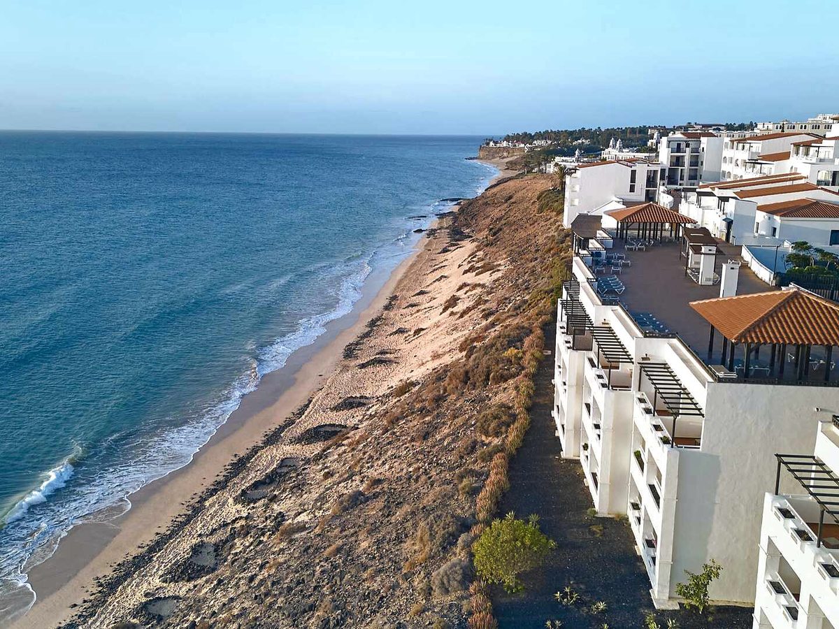 The coastline below the resort, with waves breaking on a narrow strip of sand beneath a low rocky cliff and white resort buildings along the top.