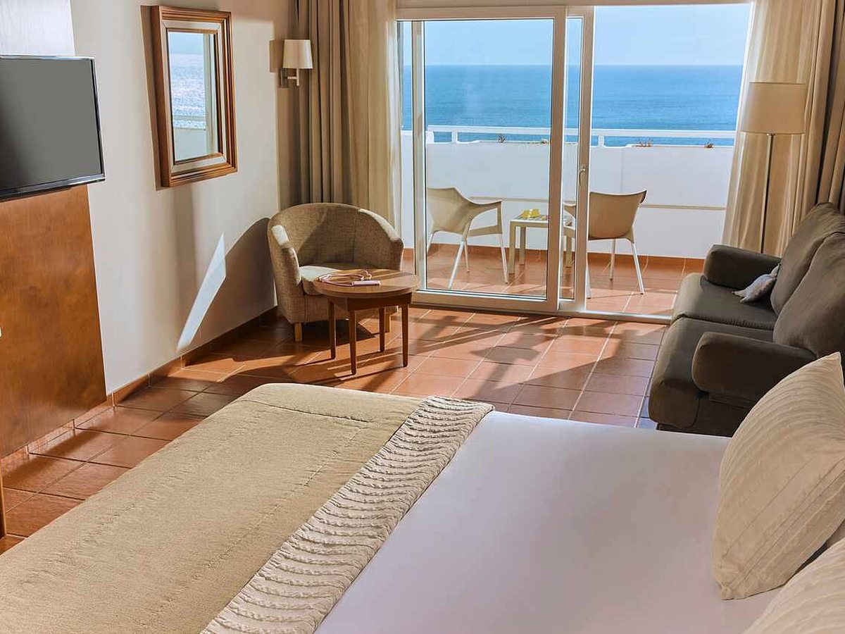 Bedroom with a sofa and armchair beside sliding glass doors onto a balcony with white chairs and a table, and the open sea beyond.