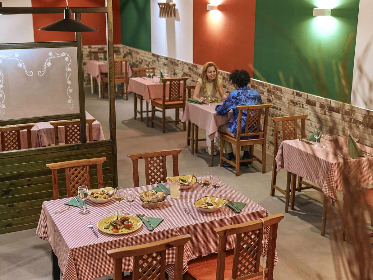 Restaurant with green, white and red walls and a brick dado, red gingham tablecloths and wooden lattice-backed chairs, with plates of pasta and bruschetta on a laid table.
