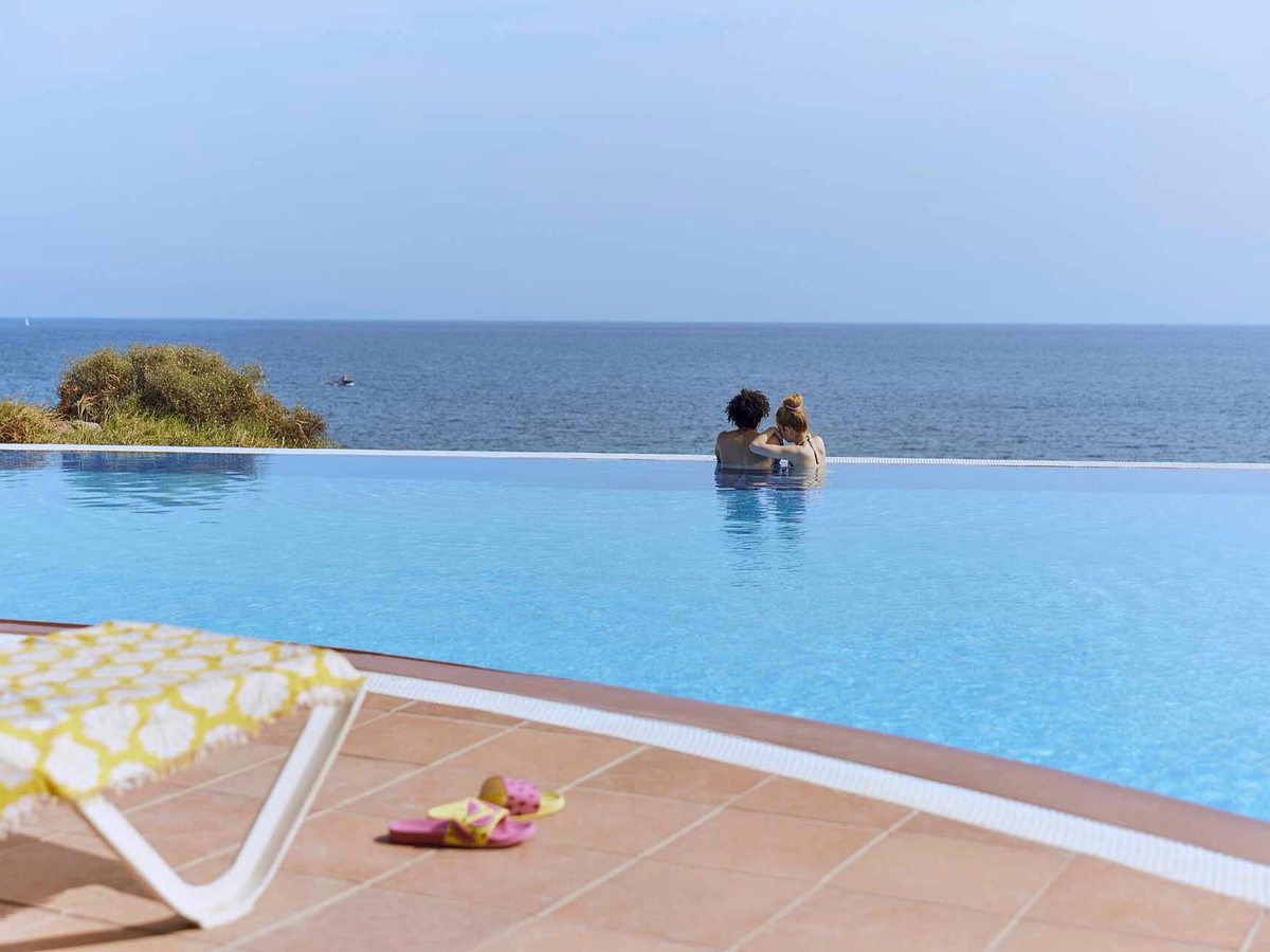 Infinity pool with its edge meeting the sea horizon, two people standing in the far end of the water and a single sun lounger on the terracotta tiled surround.