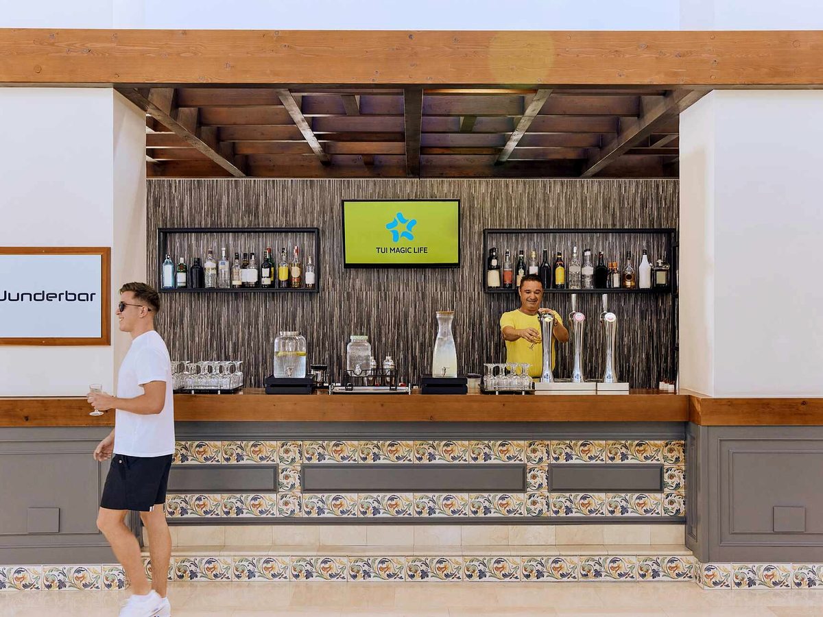 Open-fronted bar with a timber counter above patterned ceramic tiles, spirit bottles on shelves, beer taps and a barman in a yellow shirt serving a guest.
