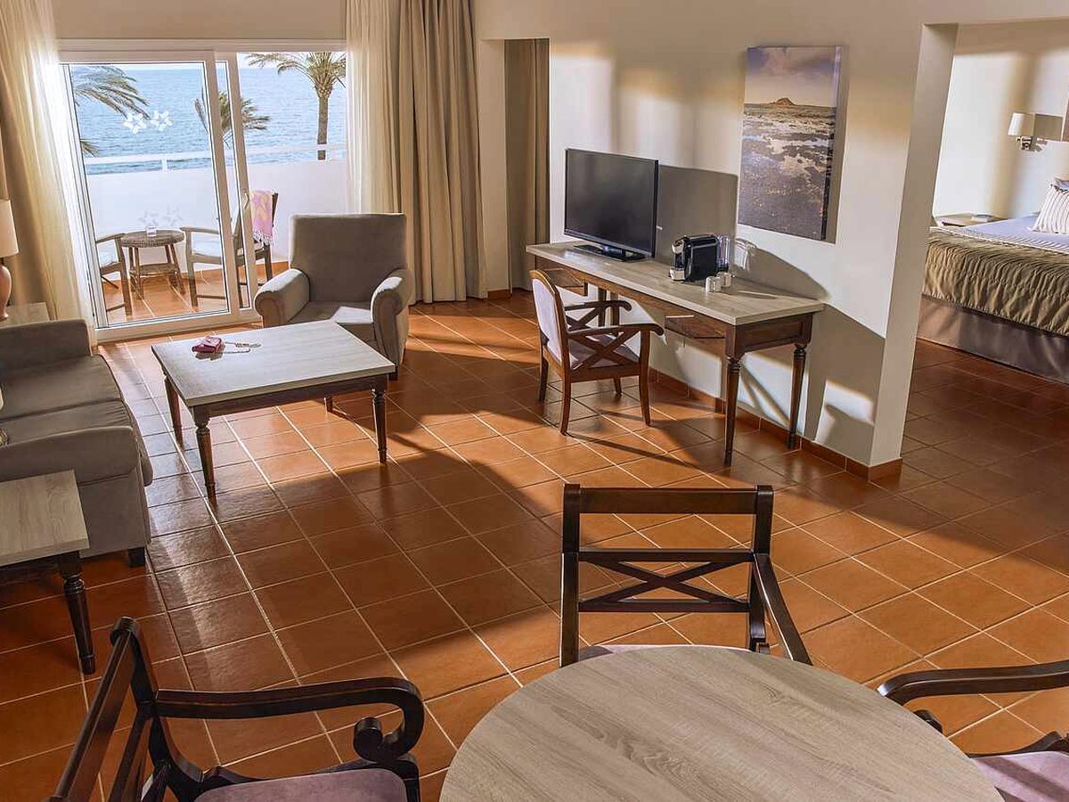 Suite living room with two sofas, a coffee table, a desk with a television and coffee machine, a dining table and chairs, the bedroom through an opening to the right and a sea-view balcony to the left.