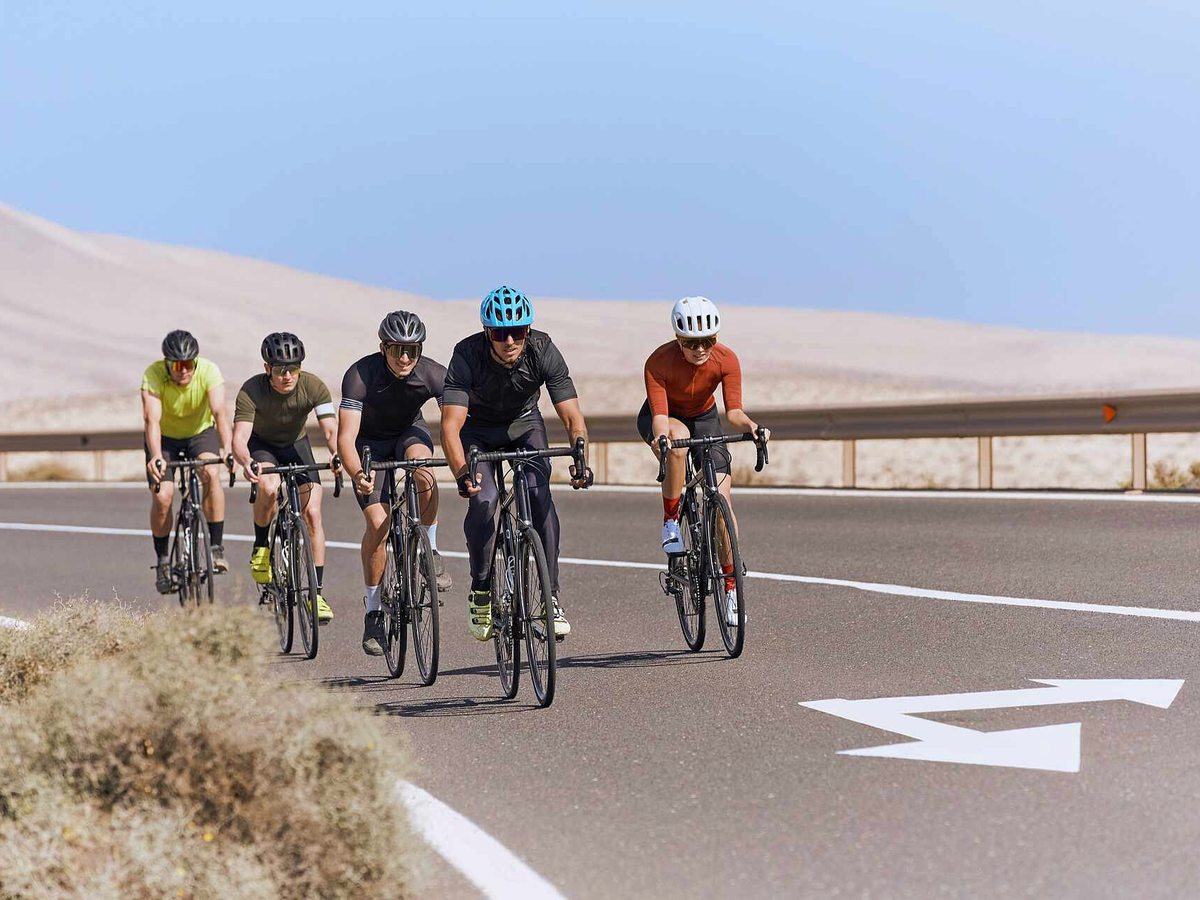 Five road cyclists riding in a group along a smooth tarmac road with white lane markings, through pale sand dunes and dry scrub.