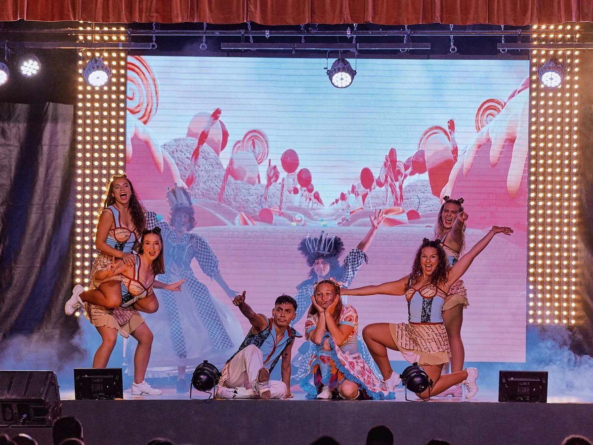 Dancers in costume posing on a lit stage in front of a large LED backdrop, with the heads of the seated audience in the foreground.