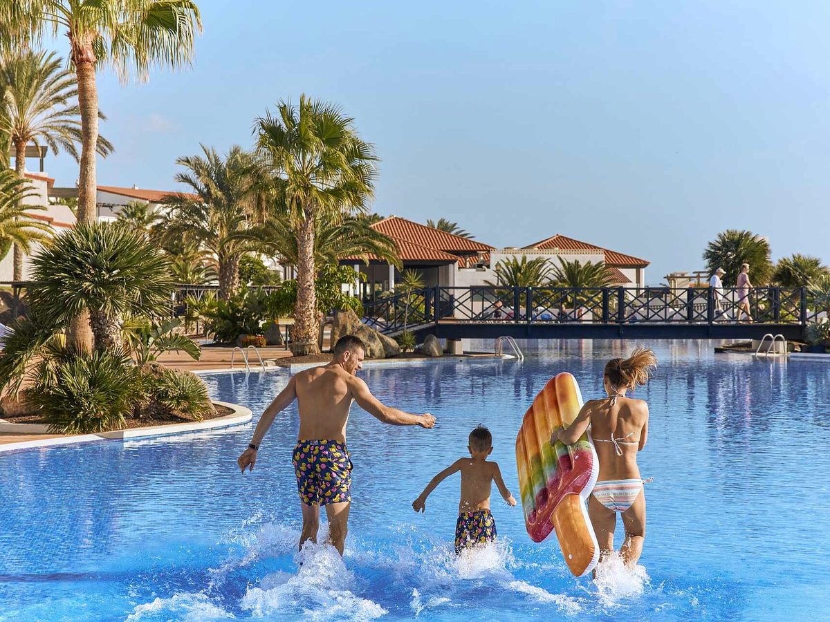 A man, a woman and a young boy walking into the shallow end of a large freeform pool, with palms, a dark timber footbridge over the water and red-roofed resort buildings behind.
