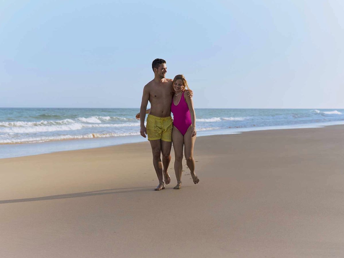 A couple walking along a wide flat sandy beach at the water's edge, with low surf breaking beside them and open sea beyond.
