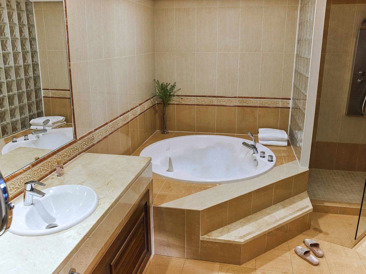 Beige-tiled bathroom with a round corner bath raised on a step, a long marble vanity with a basin and wide mirror, and a glass shower cubicle to one side.