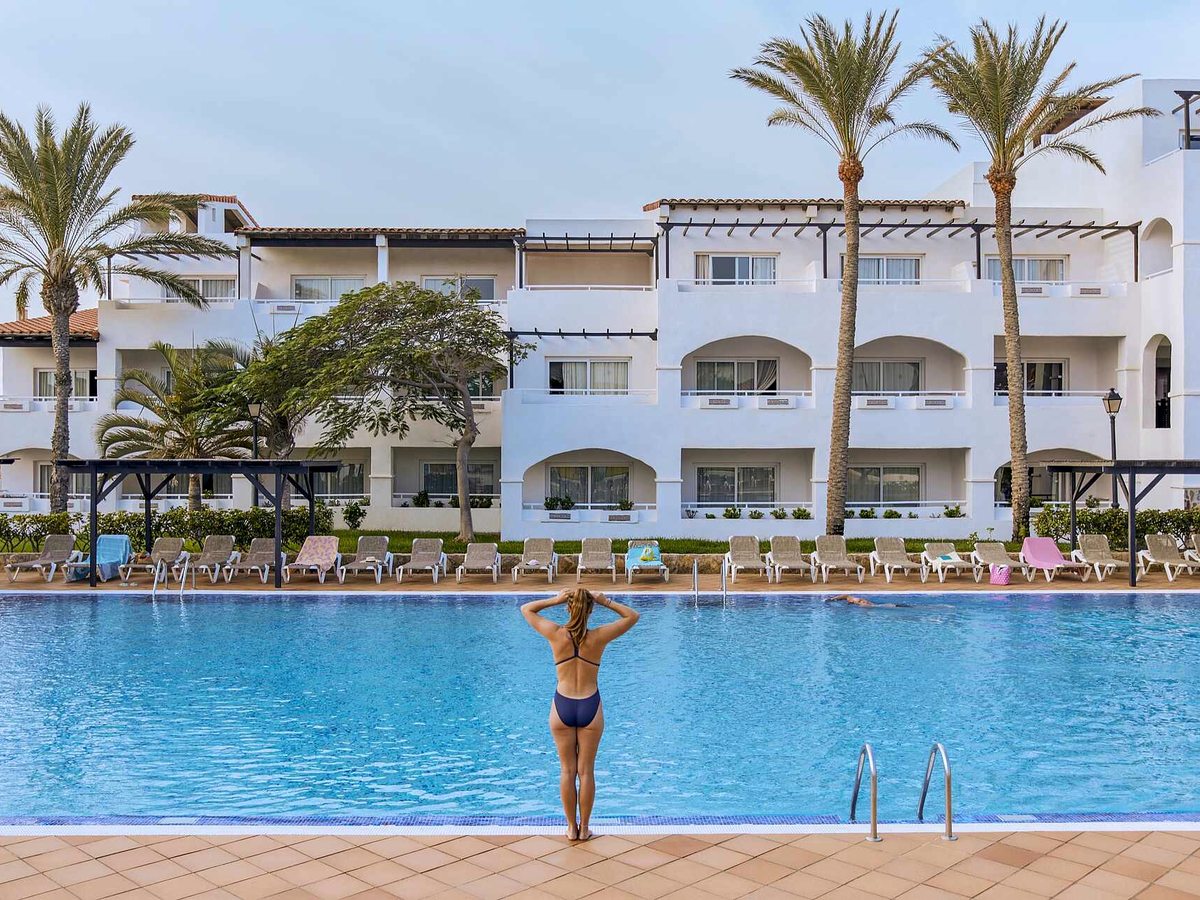 Large rectangular pool with a row of sun loungers along the tiled surround, a swimmer doing lengths, and a white block of arched balconies and palms behind.