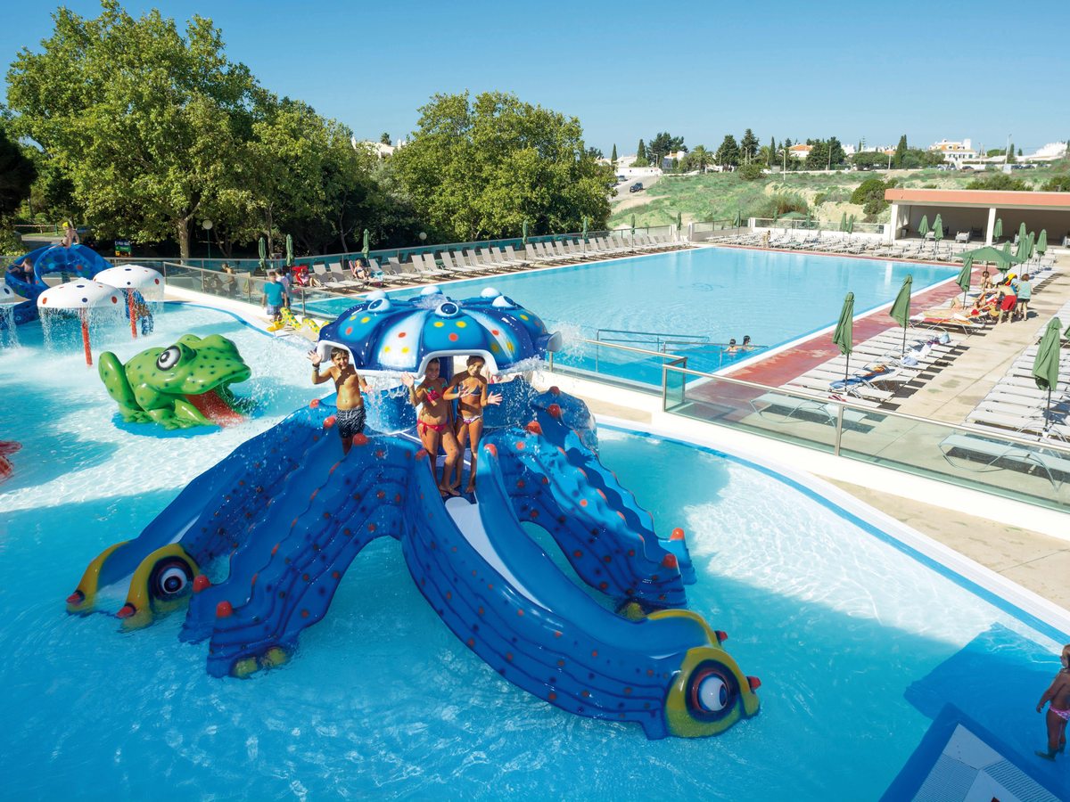 Children's splash pool with a large blue octopus-shaped slide, mushroom water sprays and a green frog feature, children playing on the slide.