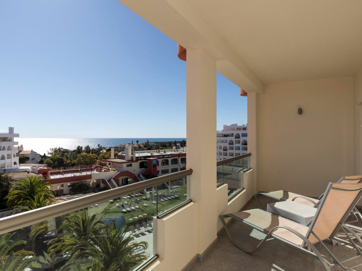 Covered balcony with two reclining chairs and a glass balustrade, above palms, resort buildings and the sea.