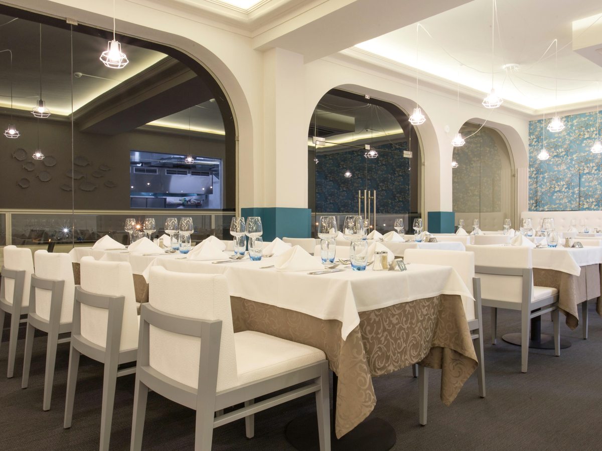 Restaurant dining room with tables laid with white cloths, wine glasses and folded napkins, white chairs, arched mirrors and pendant lights.