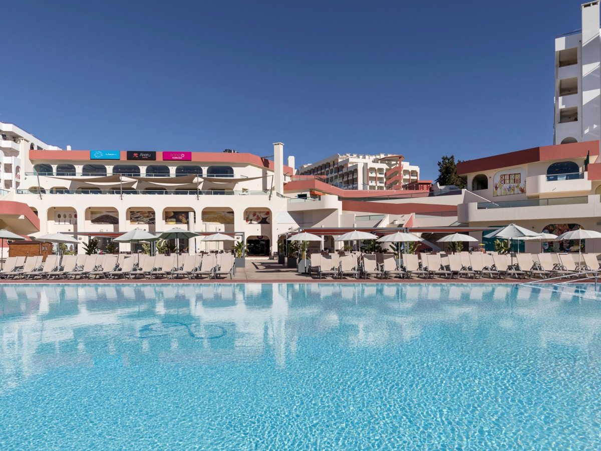 Wide blue outdoor pool with rows of sun loungers and parasols in front of a white arcaded leisure building.
