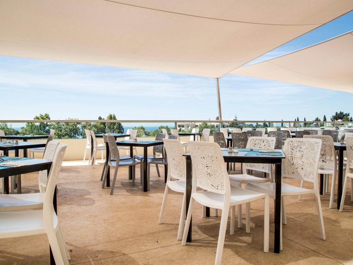 Outdoor dining terrace under white shade sails, with dark tables set for a meal, white chairs and the sea beyond the railing.
