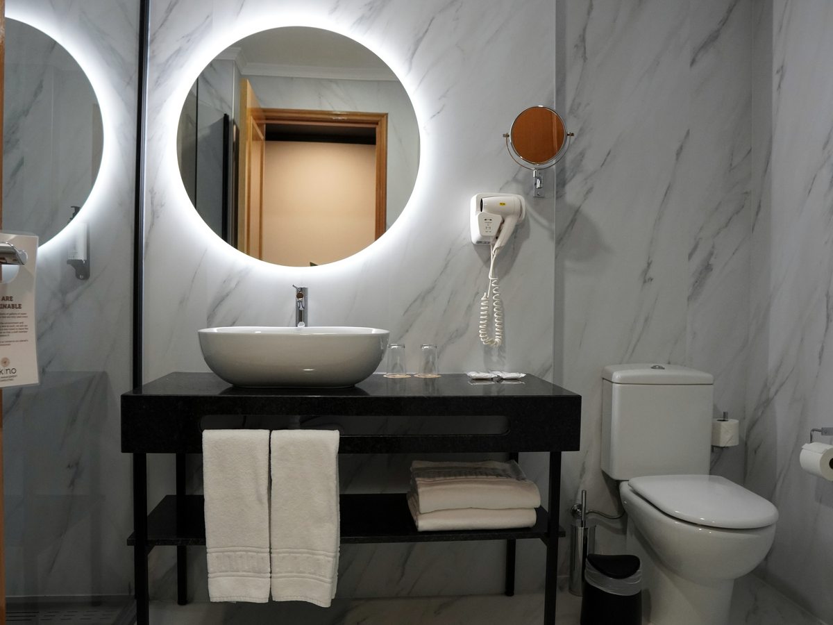 Bathroom with marble-effect walls, a vessel basin on a black vanity unit, a round backlit mirror, folded towels and a toilet.