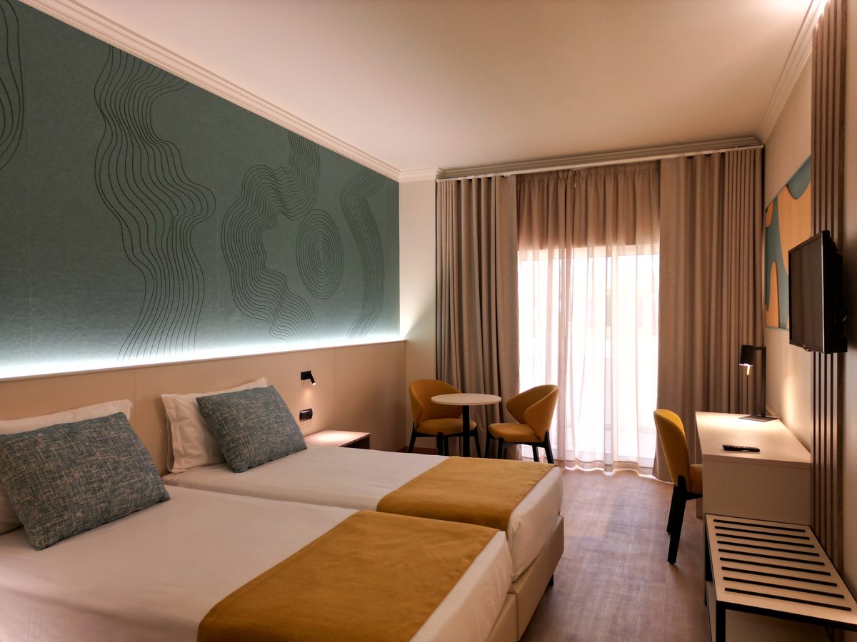 Hotel room with two single beds under mustard throws, a green patterned feature wall, two chairs and a small table, and balcony doors behind sheer curtains.