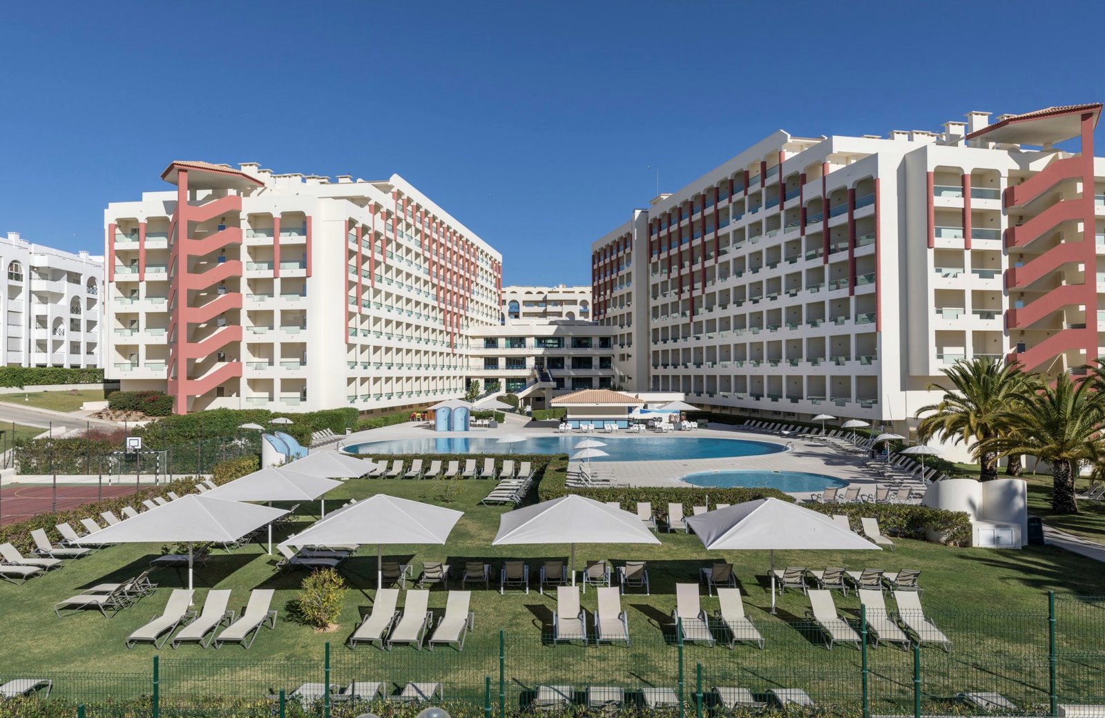 Two eight-storey white and terracotta accommodation blocks at Ukino Palmeiras Village, set around a curved blue pool with sun loungers and parasols on the lawn in front.
