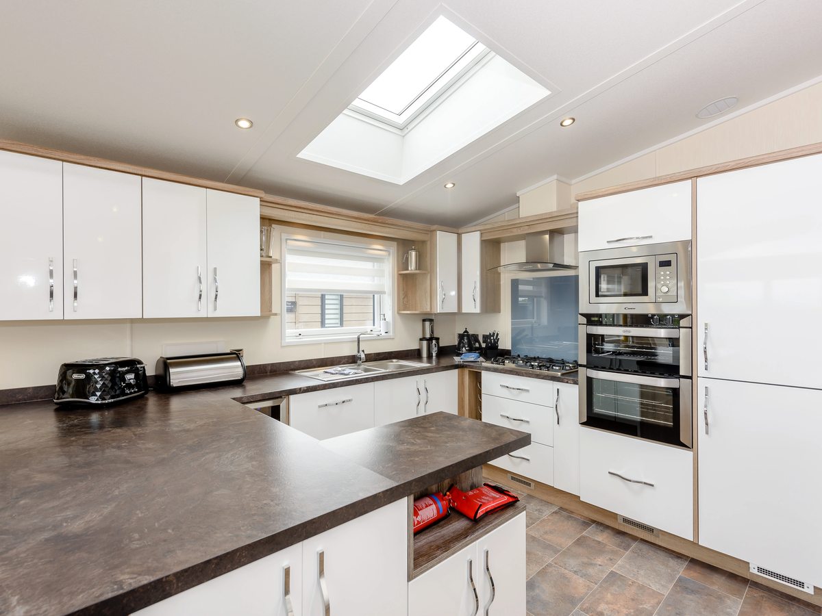 A U-shaped lodge kitchen with white cabinets, dark worktops, a gas hob, a stacked oven and microwave and a skylight above.