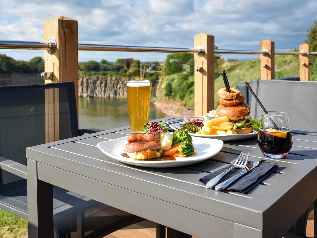 A table on the Lake House terrace set with a burger, chips, onion rings and salad, a pint of beer and a glass of wine, with the quarry lake behind.