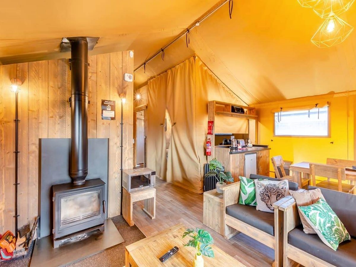The open-plan living area of a safari tent with a log-burning stove, timber-framed sofas, a coffee table and canvas walls.