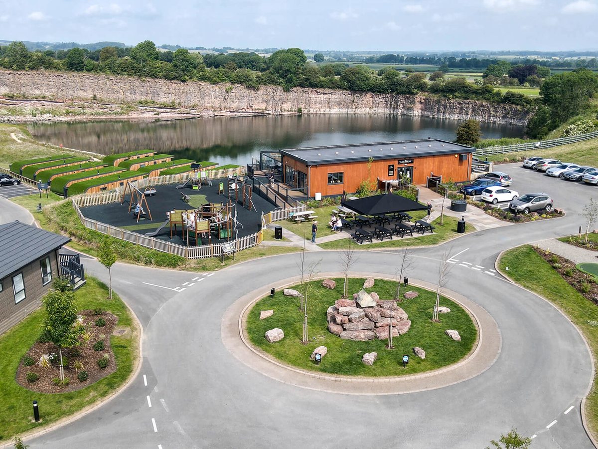 An aerial view of the Lake House restaurant beside the quarry lake, with an enclosed playground, picnic tables and a car park around a landscaped roundabout.