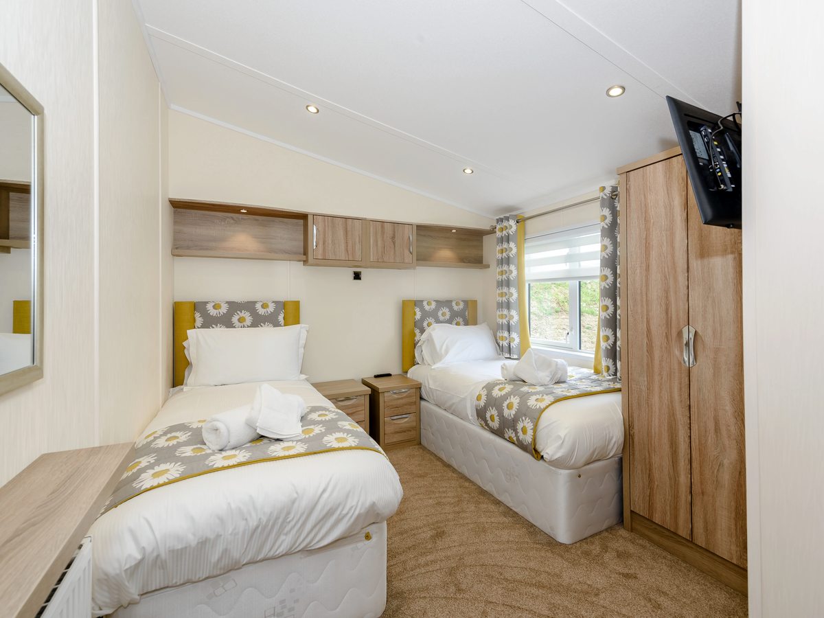 A lodge twin bedroom with two single beds, bedside tables, overhead cupboards and a full-height mirrored wardrobe.