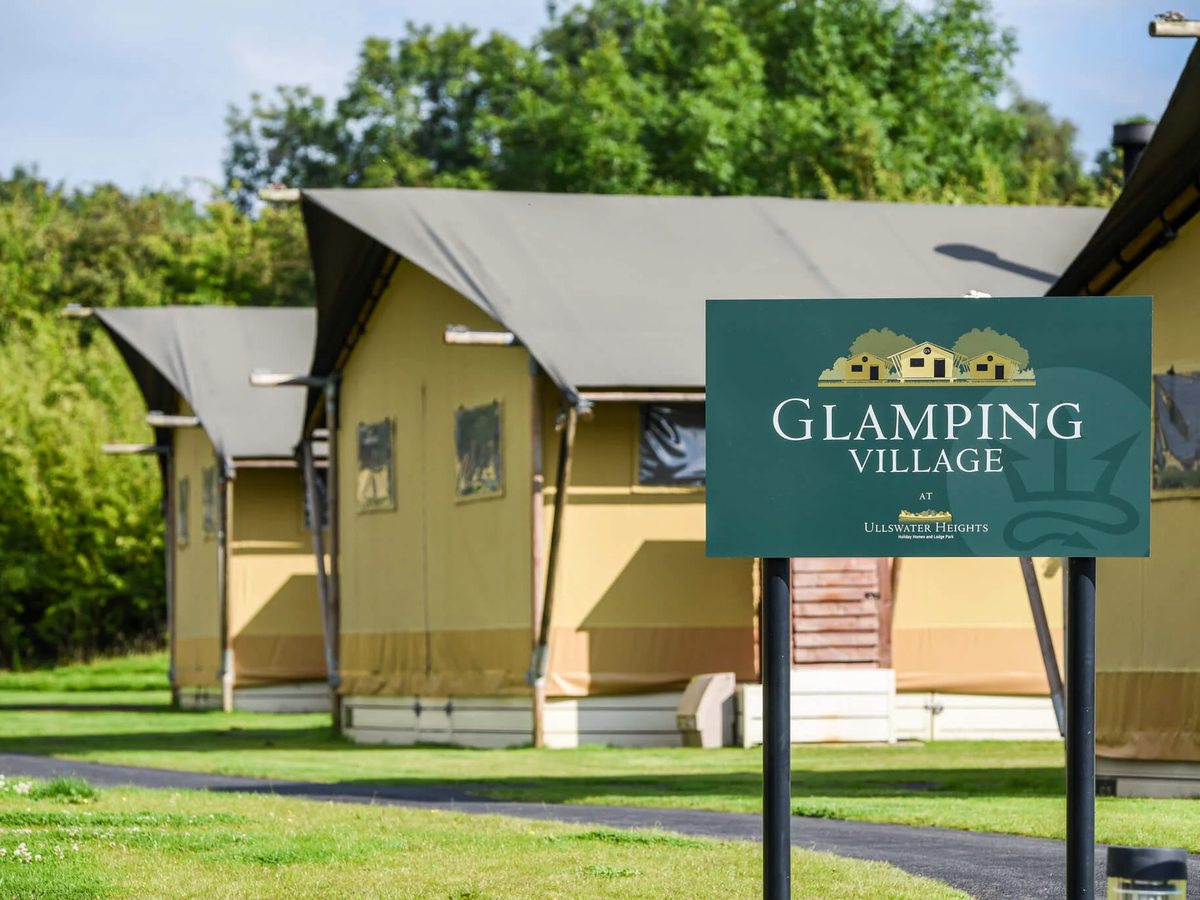 A carved Glamping Village sign on a post in front of canvas-and-timber safari tents on a mown lawn.