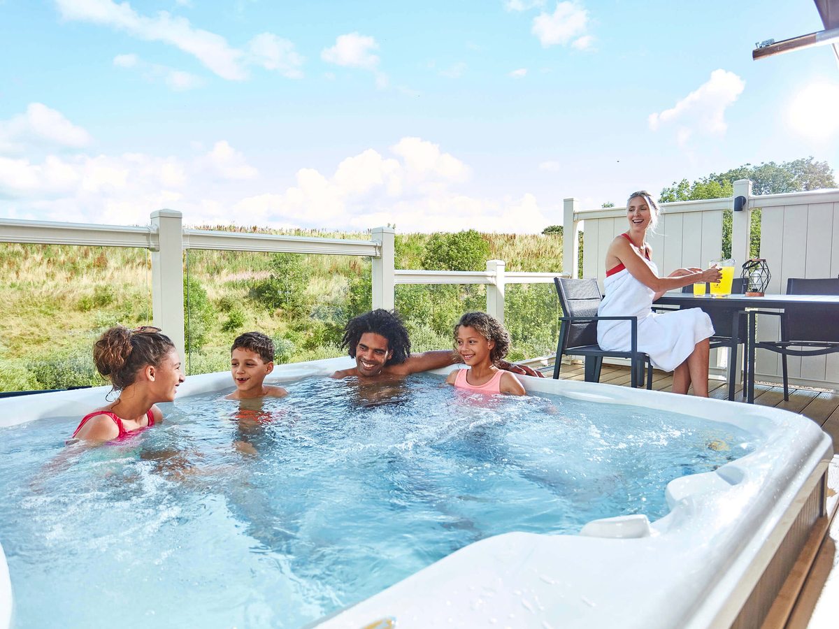 Three children and an adult sitting in a bubbling hot tub on a deck, with another adult at a table beside it and grassland behind.