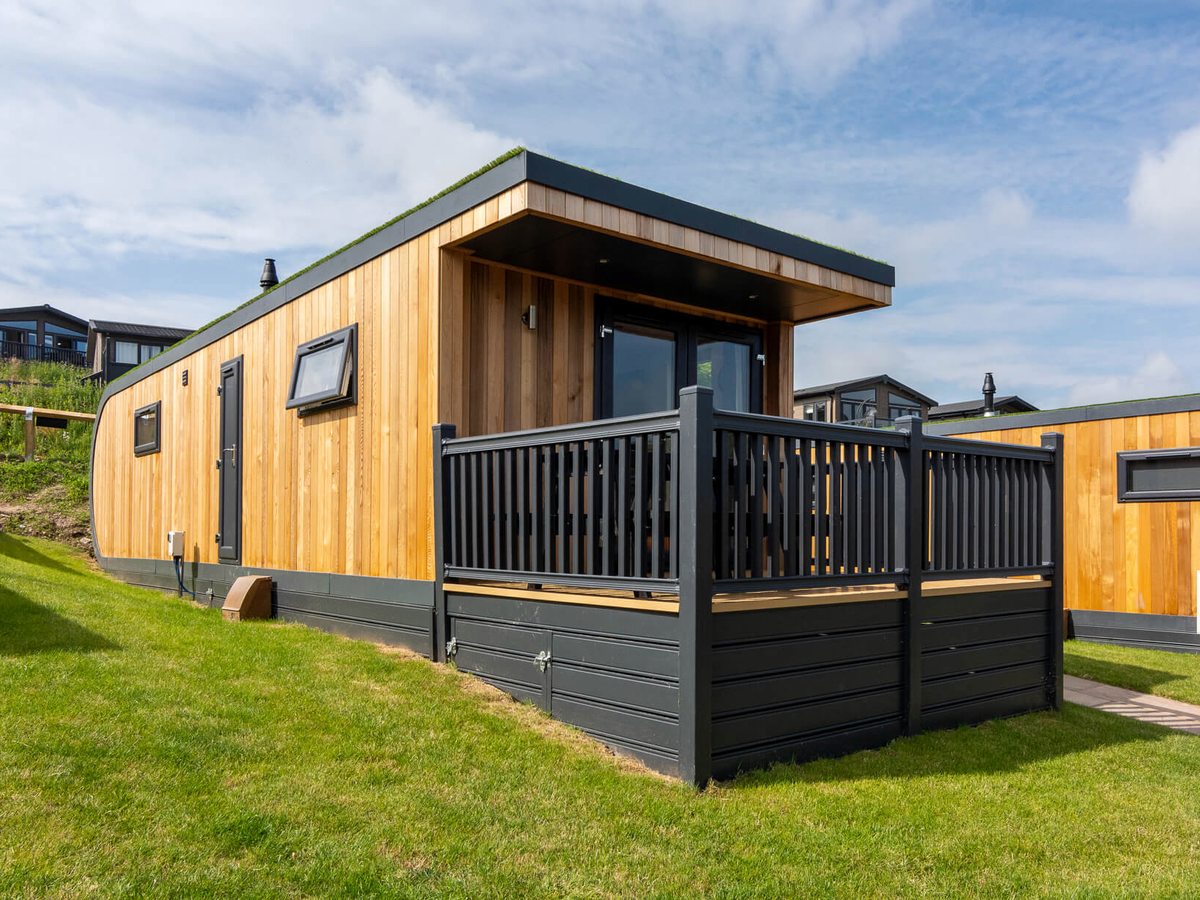 A compact timber-clad pod with a turfed green roof curving down at one end, a railed deck and glazed doors, standing on mown grass with lodges on the bank behind.
