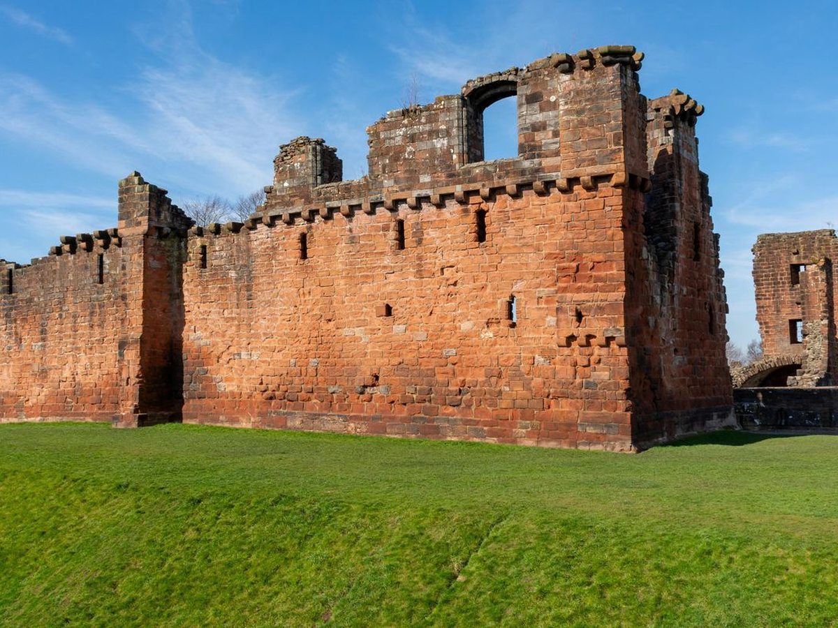 The red sandstone curtain wall and towers of Penrith Castle standing on a grassy lawn under a blue sky.