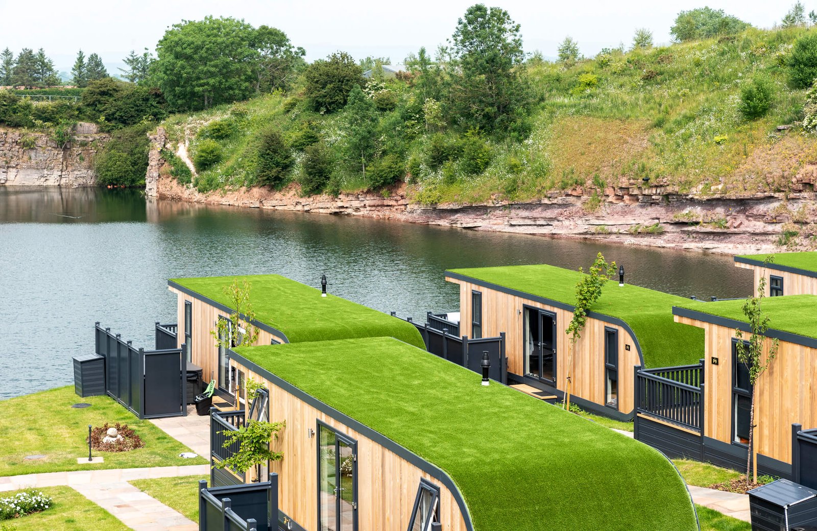 Timber lodges with living green roofs and private fenced decks lining the edge of the quarry lake at Ullswater Heights, with red sandstone quarry banks behind.