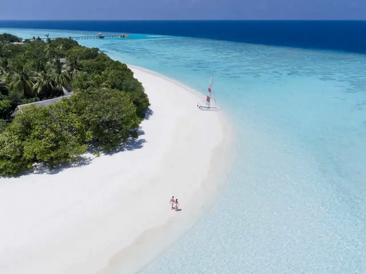 Aerial view of a curving white sand shore backed by palms, with two people walking, a small sailing catamaran on the sand and clear turquoise shallows.