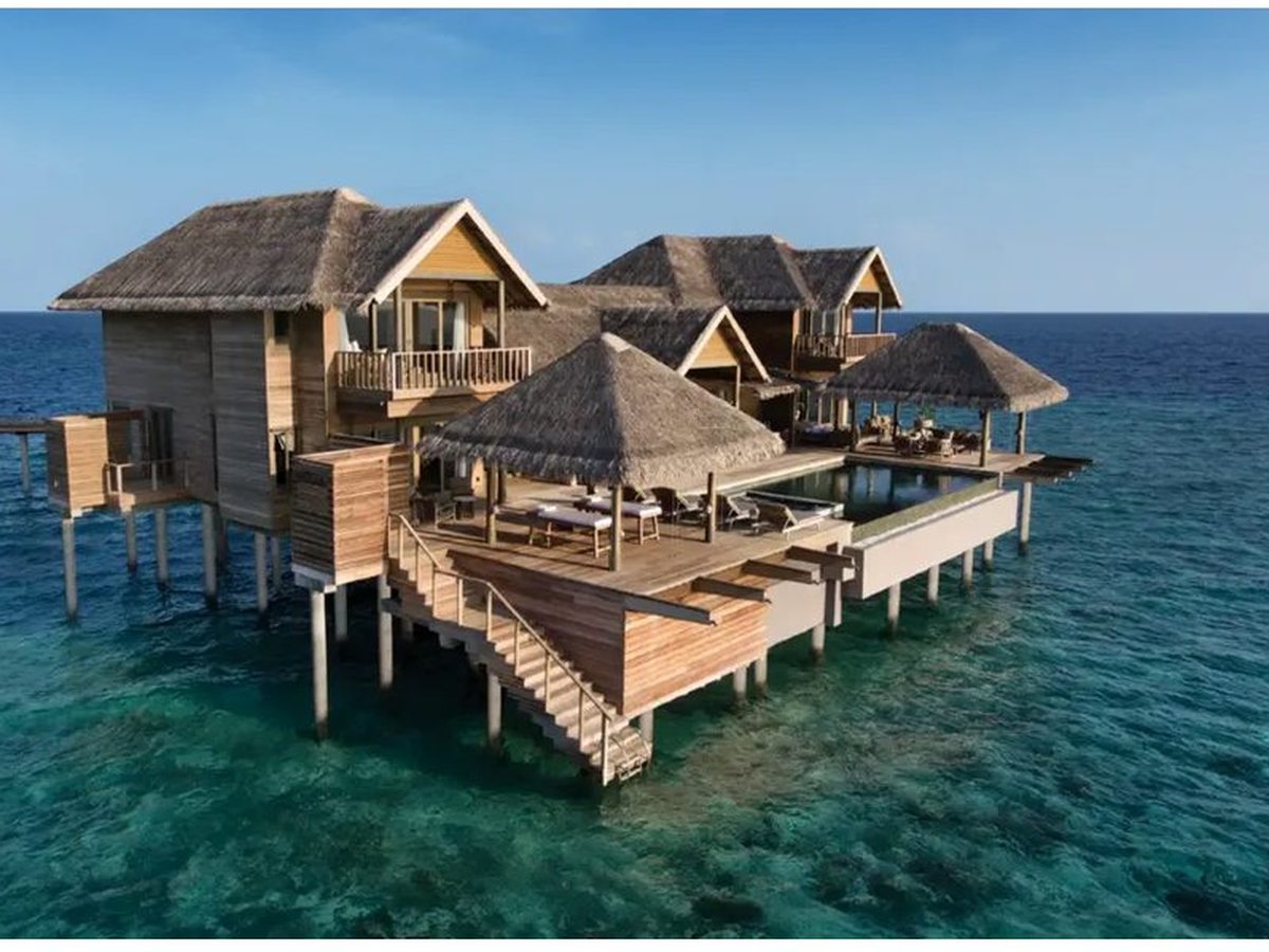 A large two-storey thatched villa on stilts above deep blue water, with a wooden sun deck, sun loungers, a pool and timber steps down to the sea.