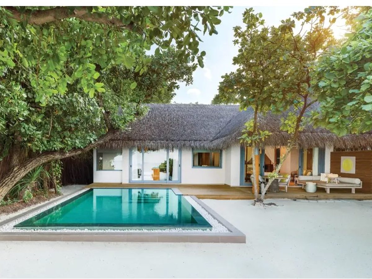 Rectangular pool set into white sand in front of a thatched-roof villa, shaded by broad-leaved trees, with a timber deck and outdoor seating alongside.