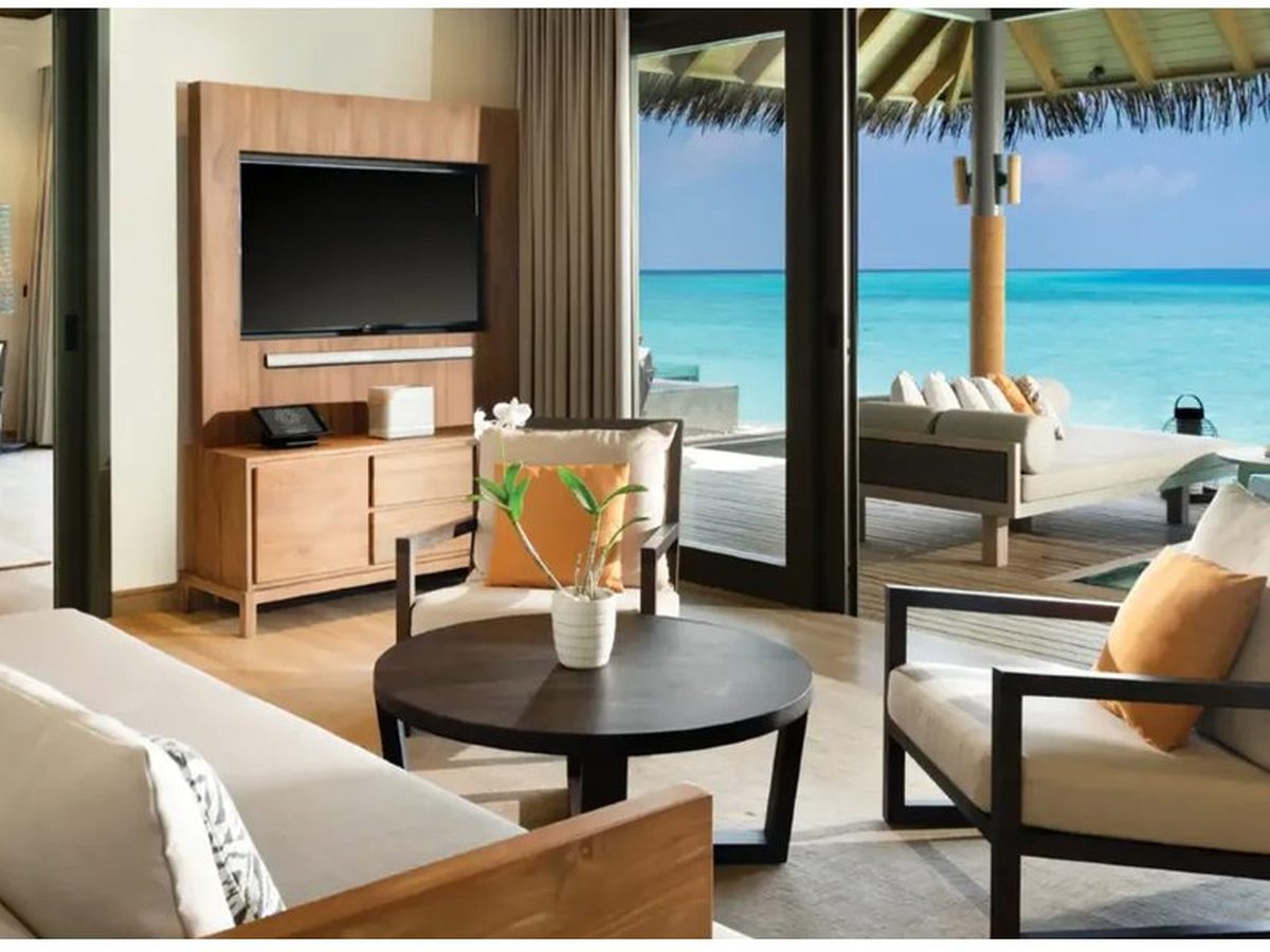 Living room with cream sofas and armchairs around a low round table and a wall-mounted television, and glass doors open onto a deck, day beds and the sea.