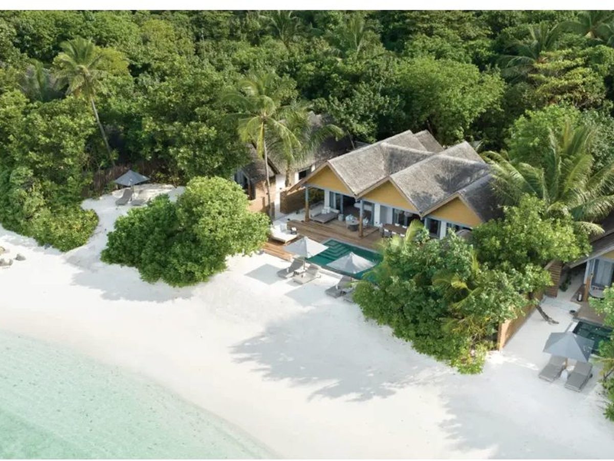 Aerial view of a thatched beach villa among trees with a timber deck, a green pool and sun loungers on the white sand, and turquoise shallows beyond.