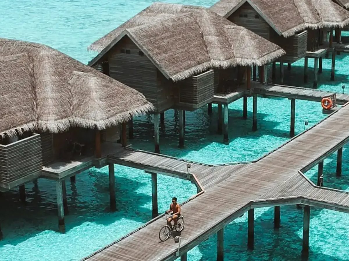 A man cycling along a wooden boardwalk above bright turquoise shallows, with thatched overwater villas on stilts on either side.