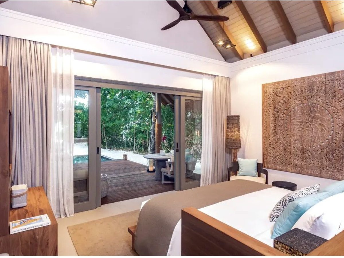 Bedroom with a king bed below a carved wooden wall panel and a pitched timber ceiling, with sliding doors open onto a deck, a small pool and trees.