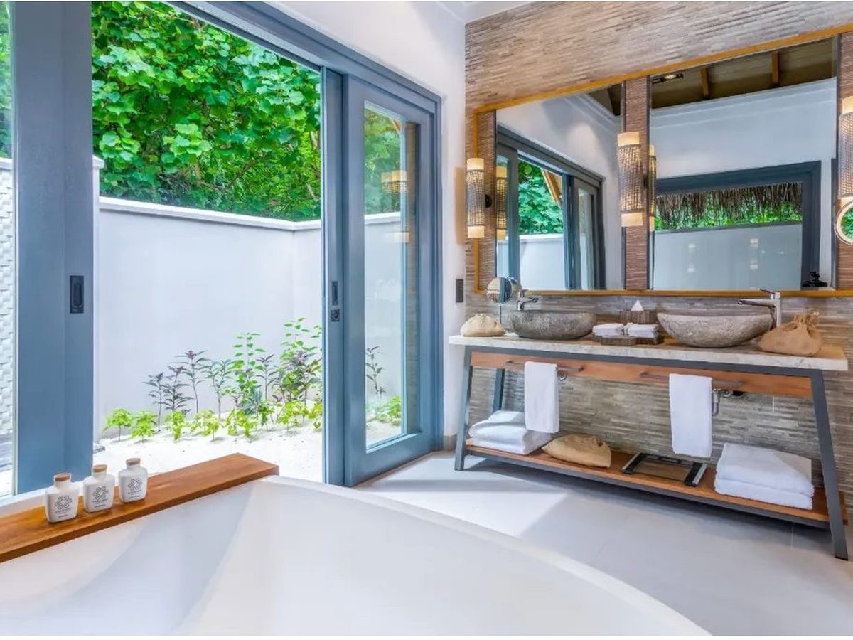 Bathroom with two stone basins on a timber vanity below a wide mirror, and a bath beside sliding doors onto a small walled courtyard planted with herbs.