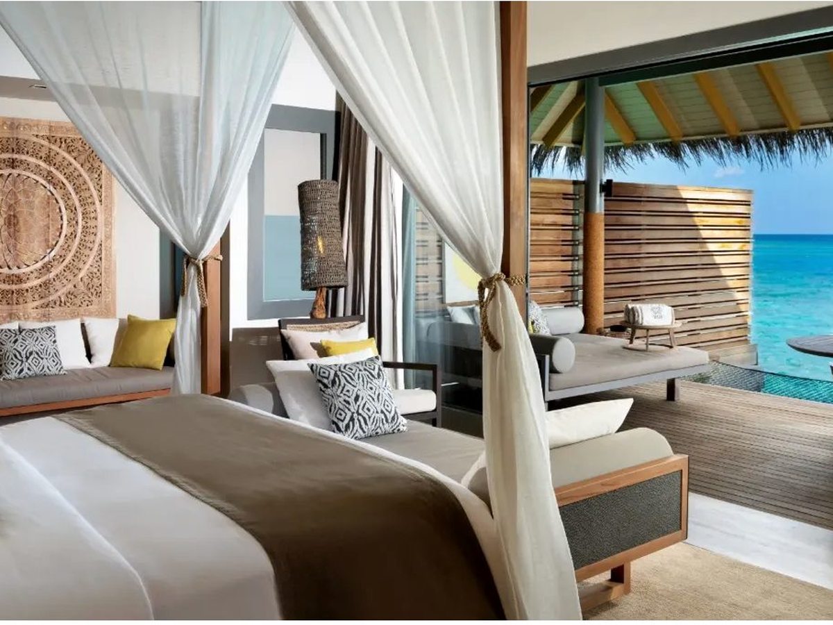 Bedroom with a white four-poster canopy bed and a daybed, opening through glass doors onto a shaded deck, a plunge pool and the turquoise lagoon.