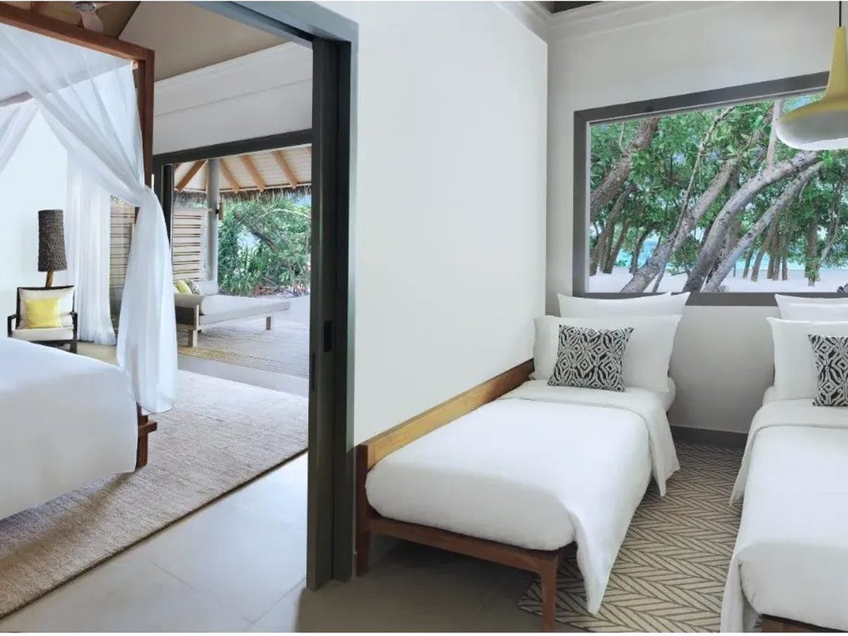 Two single beds with white linen and patterned cushions beneath a wide window onto the trees, with the main bedroom and its canopy bed through an open sliding door.