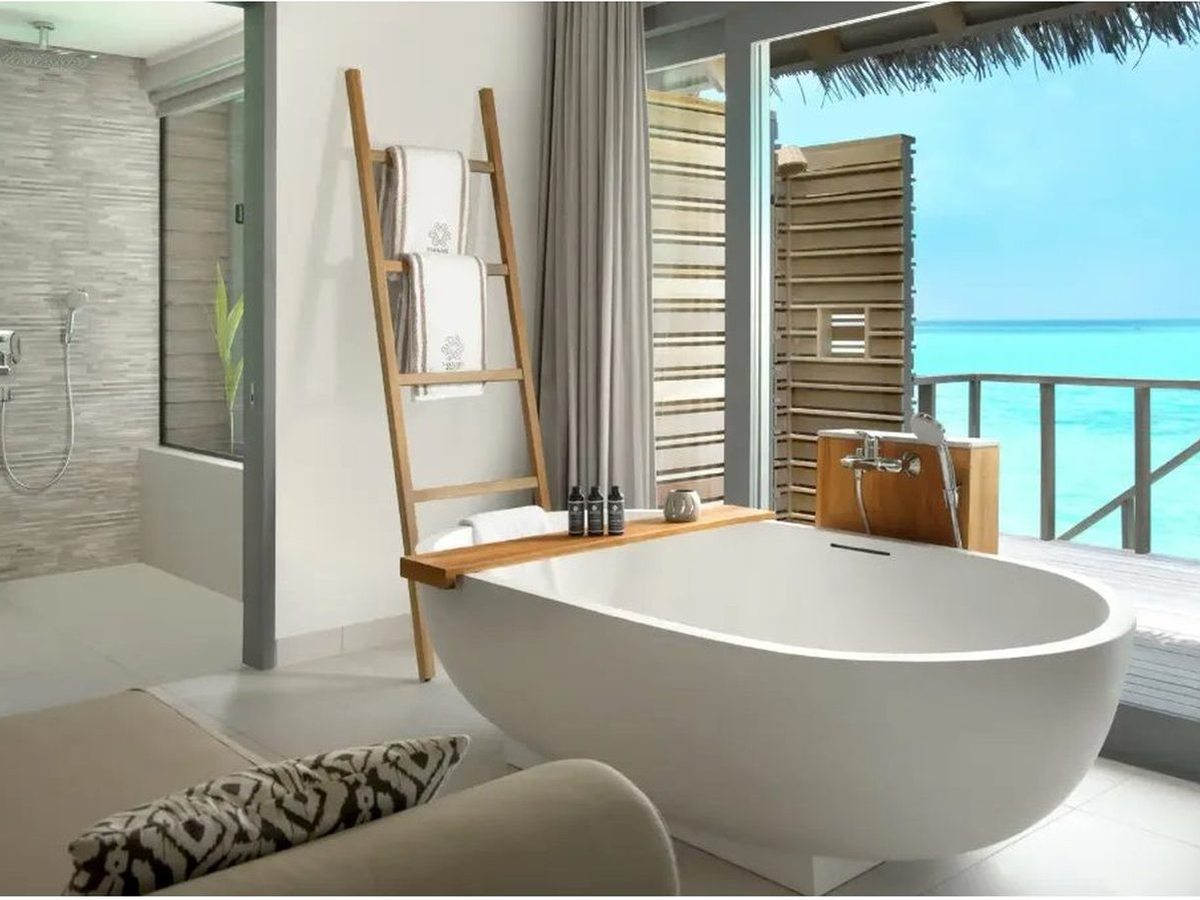 Bathroom with a freestanding oval bath and a timber towel ladder beside a walk-in shower, and glass doors onto a deck above turquoise water.