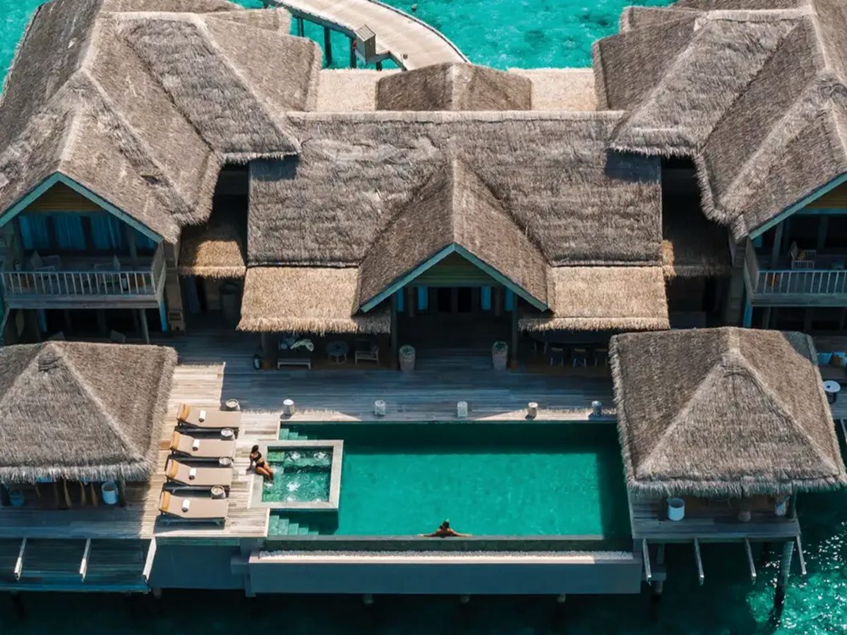 Overhead view of a long pool and whirlpool on the sun deck of a large thatched overwater residence, with sun loungers along one side.