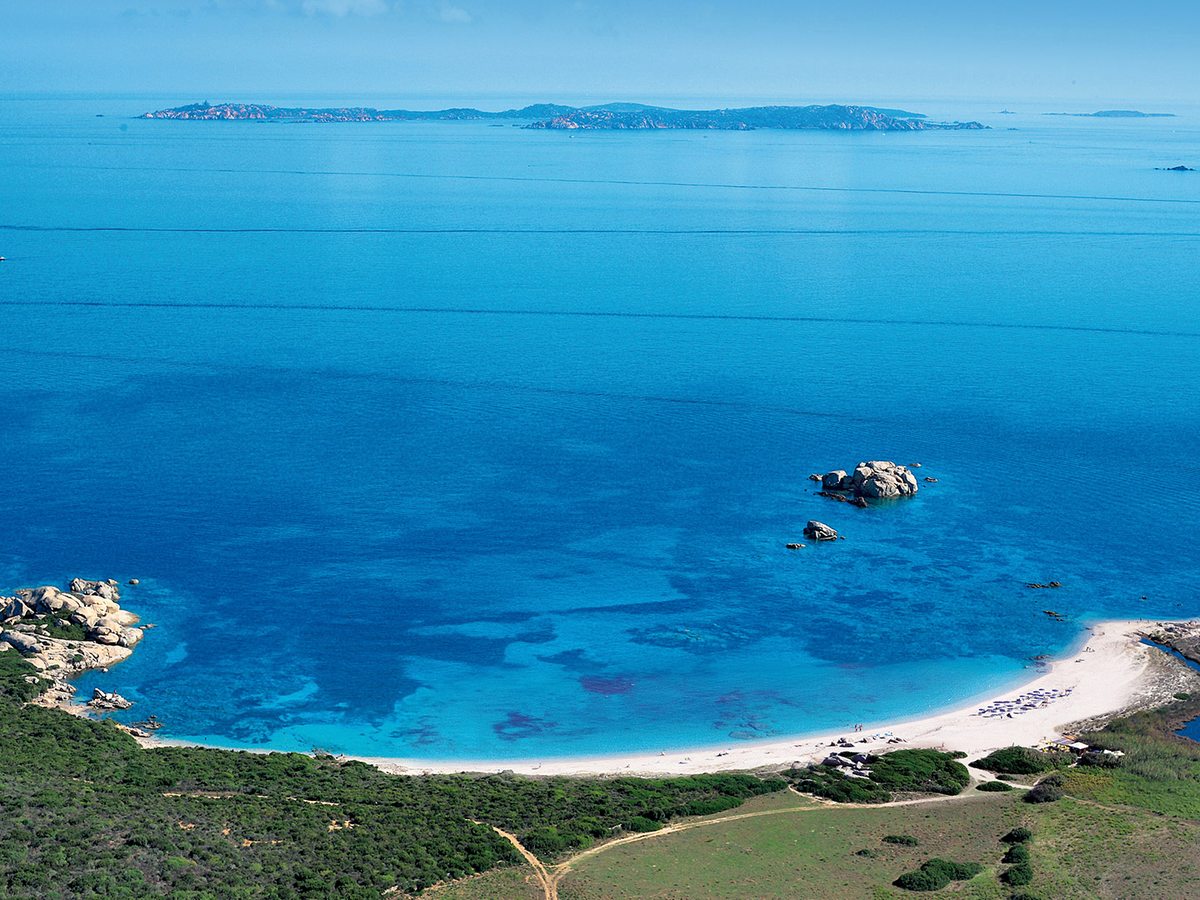 Elevated view of a long white sandy beach curving around a bay, with clear turquoise shallows deepening to blue and a low island on the horizon.