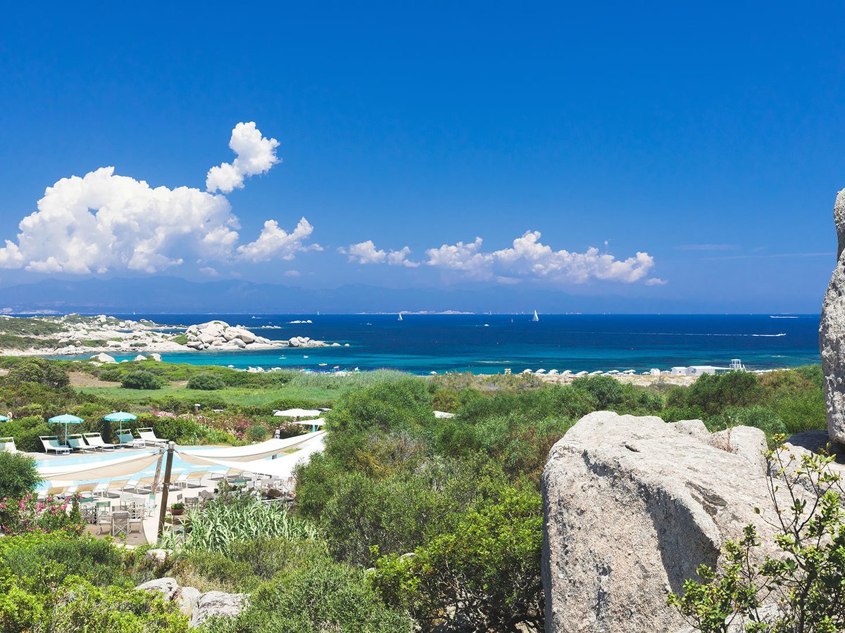 White shade sails, sun loungers and parasols on a terrace in the foreground, with green scrub, a sandy beach and blue sea under a bright sky beyond.