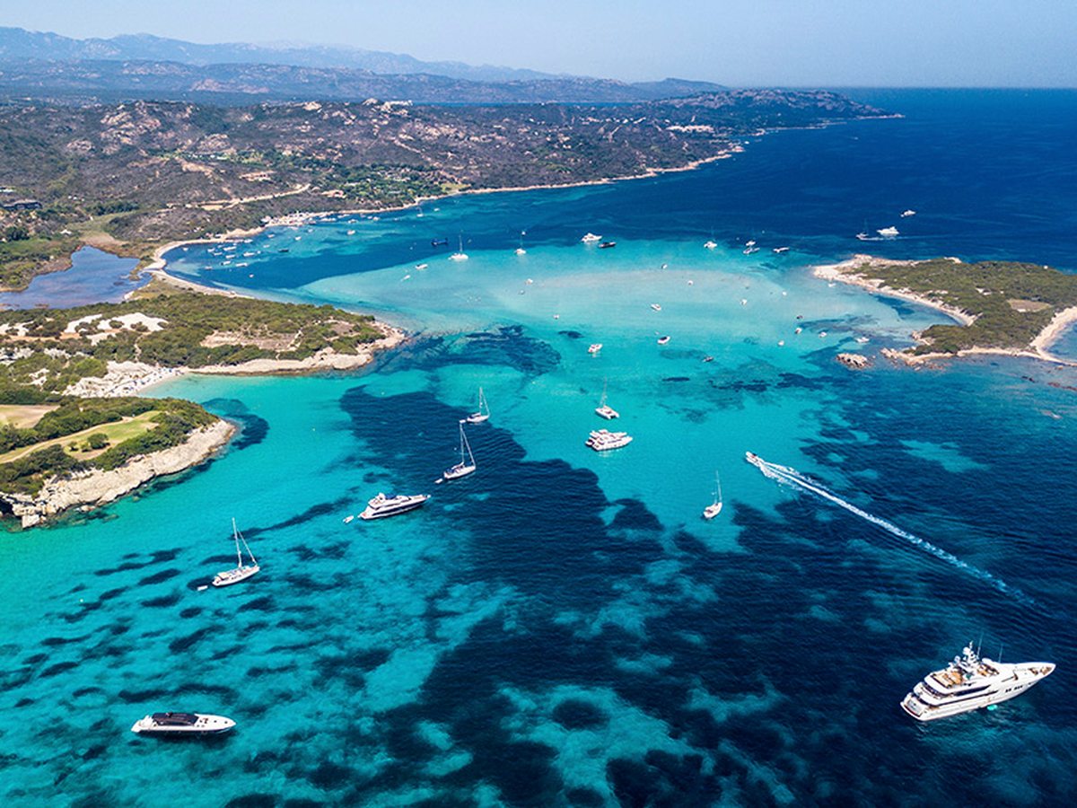 Aerial view of a wide turquoise bay with yachts and motorboats anchored over pale sand, wooded headlands on either side and hills behind.