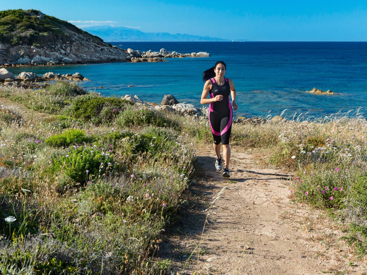 A woman running along a narrow sandy path through low green and yellow scrub, with granite boulders and the blue sea behind.