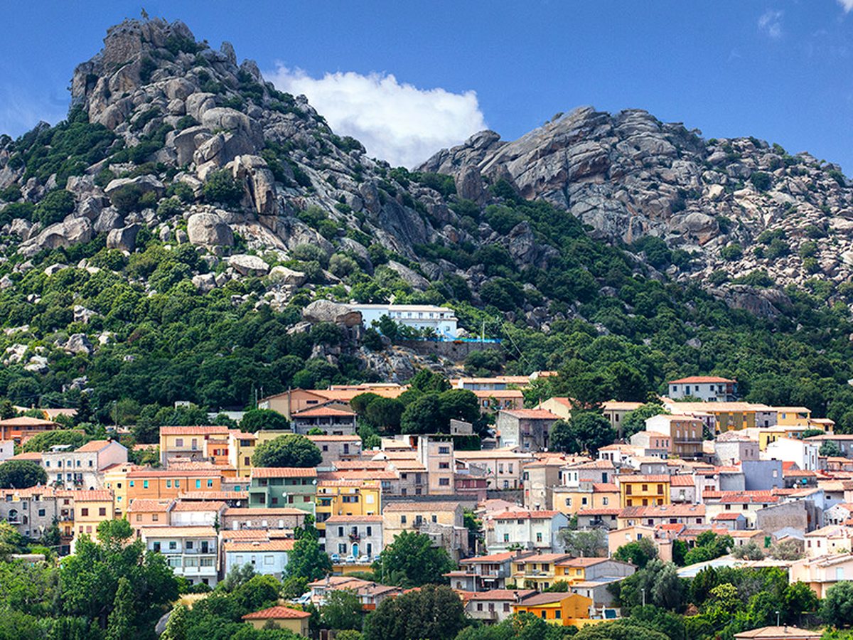 A village of pastel-painted houses on a slope below a jagged granite mountain, with trees around the rooftops.