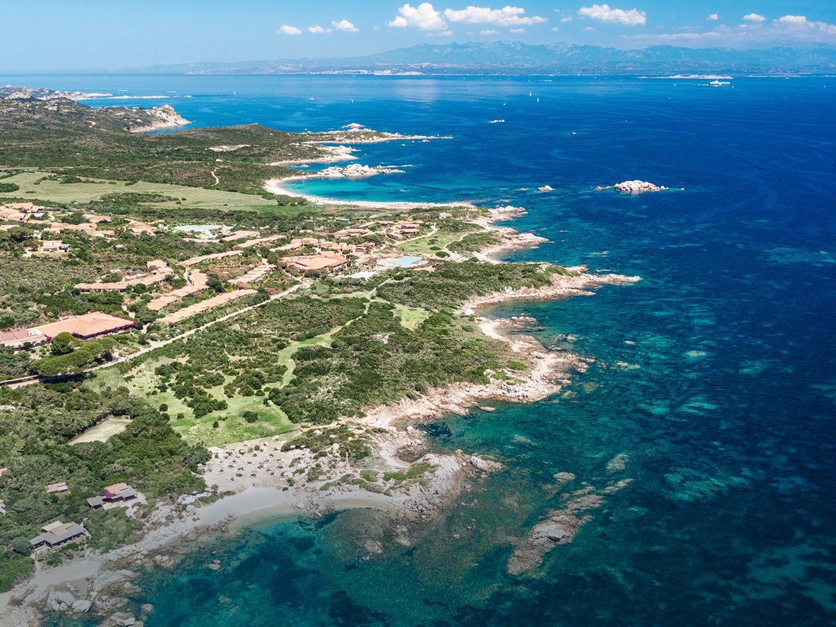 Aerial view of low terracotta-roofed buildings, pools and green lawns set among scrub along a rocky coastline indented with small sandy coves, with deep blue sea and distant hills beyond.